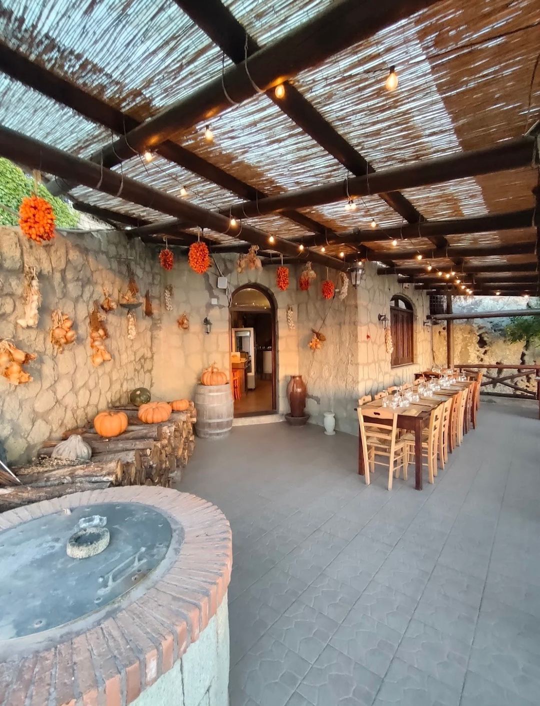 A rustic outdoor dining area under a wooden pergola with string lights, decorated with autumn pumpkins and hanging dried peppers, featuring a brick fire pit in the foreground.
