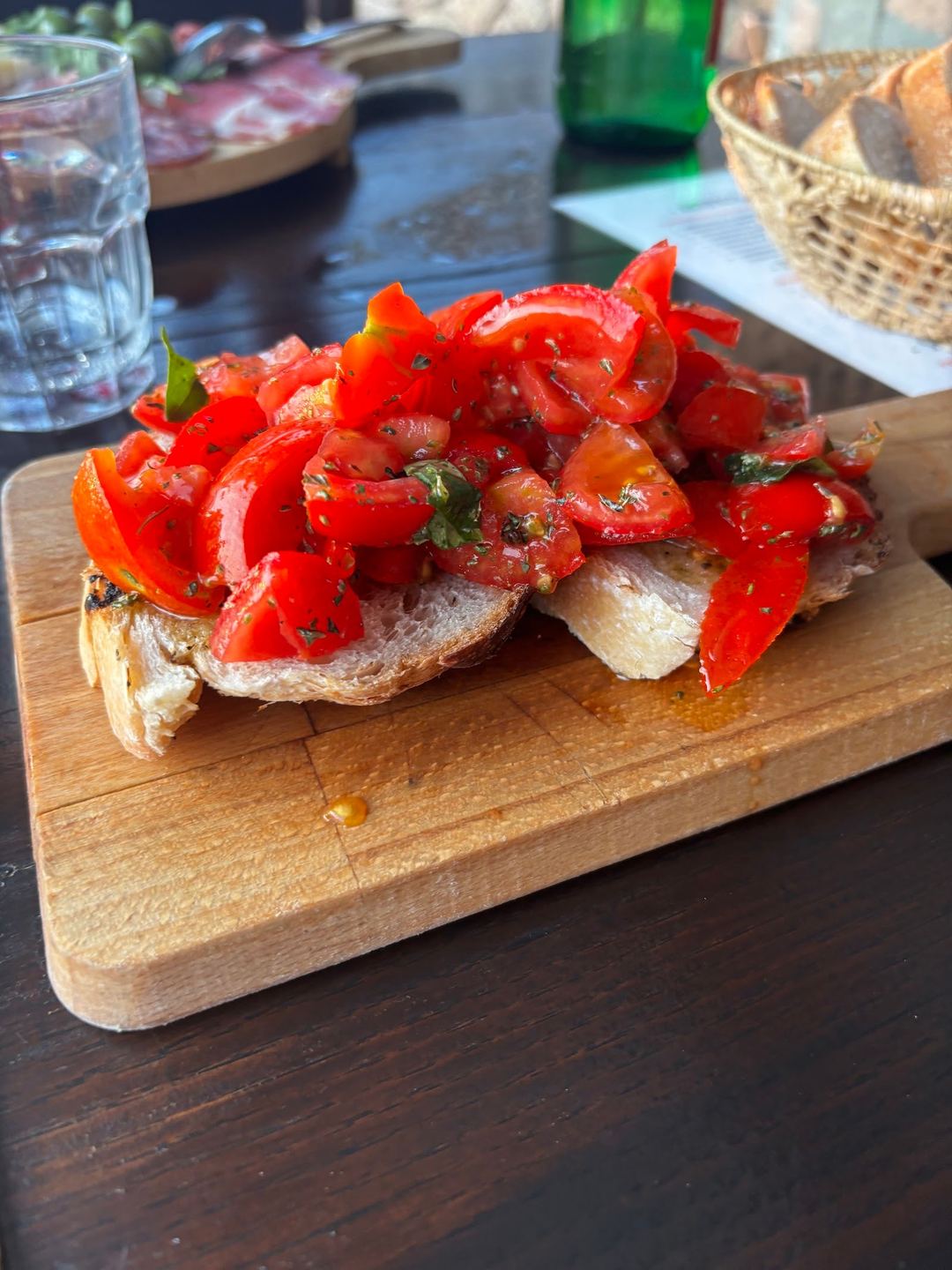 Toasted bread topped with fresh roasted red peppers and herbs, served on a wooden board with water and bread basket in the background.