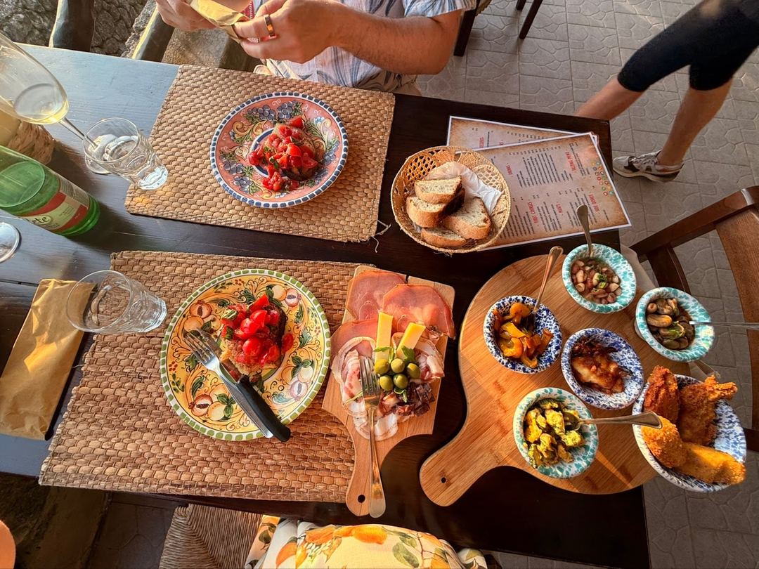 An overhead view of an agriturismo dining table featuring colorful ceramic plates with roasted vegetables, cured meats, fresh bread, and traditional Italian dishes served during a group meal.