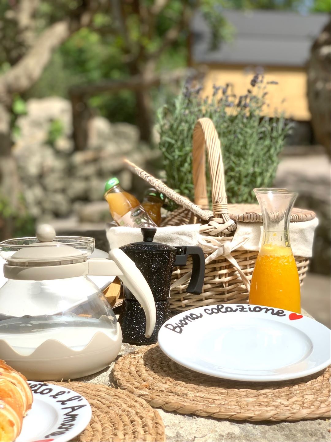 An outdoor breakfast table setting with personalized plates, fresh orange juice, pastries, and traditional Italian tableware arranged in a sun-dappled Mediterranean garden courtyard.