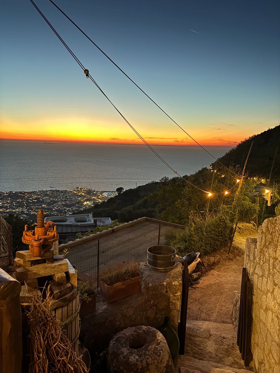 Sunset view over the Mediterranean Sea from the agriturismo's hillside property with rustic farm equipment and village lights below.